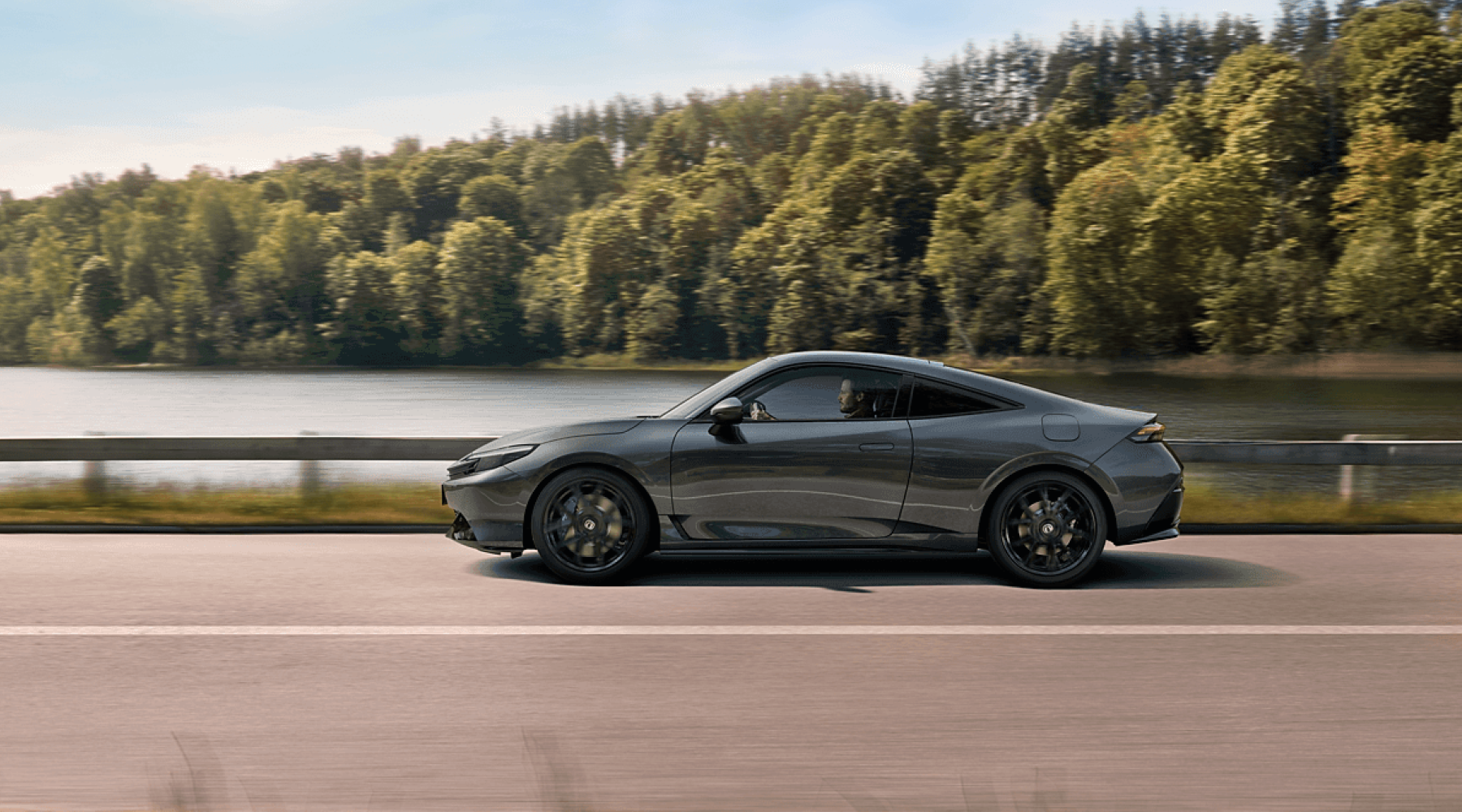 A sleek gray Honda Civic coupe driving on a road alongside a lake. The car is in motion, with dense green forest in the background and the lake visible to the left. The scene depicts a serene natural setting contrasting with the modern, sporty design of the vehicle.